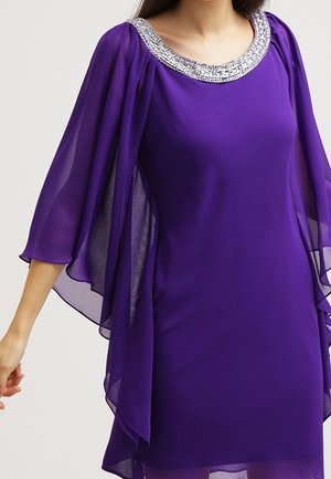 Kobieta w fioletowej sukience z szyfonu z luźnymi rękawami w formie peleryny oraz srebrnym, obszytym koralikami dekoltem, stojąca na jednolitym tle.