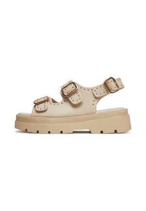 Sandalias beige con suelas gruesas, múltiples correas con hebillas y detalles con tachuelas. Hechas de material suave con tacón plano.