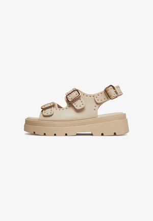 Sandali beige con suole spesse, più cinturini con fibbie e dettagli borchiati. Realizzati in materiale liscio con tacco basso.