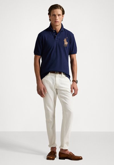 Marineblauwe polo met kraag en geborduurd ontwerp, gecombineerd met witte broeken en bruine instappers. Gladde stof met een klassieke pasvorm.