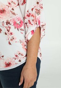 Blusa floreale con motivi di fiori rosa e rossi su sfondo bianco, con maniche arrotolate e dettaglio con bottoni.