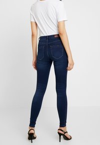 Jeans skinny azul oscuro de cintura alta, con bolsillos traseros y un sutil desgaste. Combinados con una camiseta blanca básica y tacones negros.