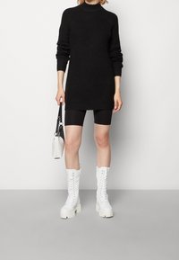 Robe-sweater noire en tricot avec un col montant, associée à des shorts de vélo noirs et des bottes blanches lacées jusqu'aux genoux. Petit sac à main blanc.