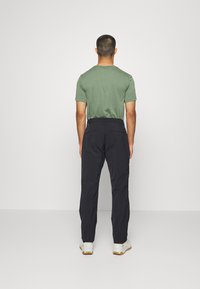 Men's outfit featuring a green short-sleeve t-shirt and black trousers with a belt, viewed from the back. Sneakers are visible.