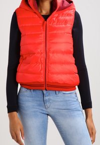 Gilet imbottito rosso con cappuccio, dotato di chiusura frontale con zip e orlo a coste. Indossato sopra una maglietta a maniche lunghe blu navy, abbinato a jeans azzurri.