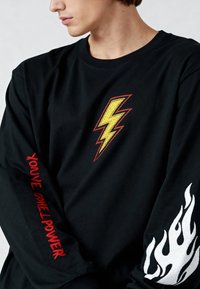 Man draagt een zwart shirt met lange mouwen met een gele bliksemschicht op de borst, rode tekst "You’ve Got The Power" op de mouw, en een wit vlammenontwerp op de andere mouw.