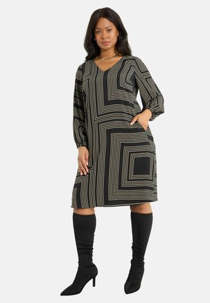 Schwarzes Kleid mit cremfarbenen geometrischen Linien, langen Puffärmeln, V-Ausschnitt und seitlichen Taschen. Getragen mit schwarzen Stiefeln mit hohen Absätzen bis zur Oberschenkel.