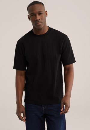 Homme portant un t-shirt noir à manches courtes côtelé et un jean bleu foncé, debout devant un fond clair uni.