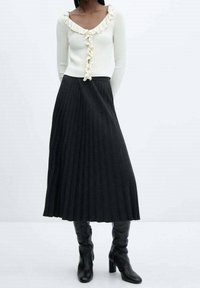 Suéter blanco acanalado con escote con volantes combinado con una falda midi plisada negra. La modelo lleva botas negras hasta la rodilla. Diseño sencillo, sin estampados.