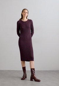 Šaty až ke kolenům, s džerzejovým vzorem v hluboké burgundské barvě, s dlouhými rukávy a kulatým výstřihem. Stylizováno s lesklými hnědými botami na středním podpatku.