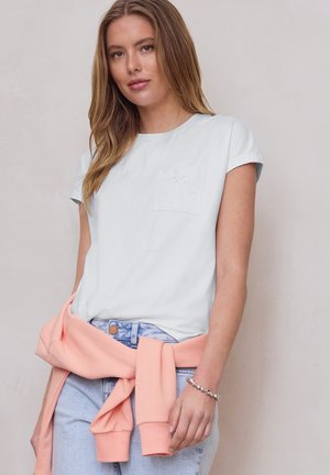 Junge Frau mit langen Haaren trägt ein weißes Kurzarmshirt, hellblaue Jeans und einen pfirsichfarbenen Pullover, der um ihre Taille gebunden ist.