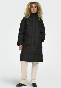 Manteau rembourré noir avec col montant, fermeture éclair et cordon de serrage. Présente une silhouette ample et des coutures visibles. Porté par-dessus un pantalon crème.