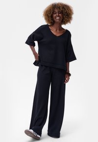 Zwarte gebreide top met een V-hals en wijde mouwen, gecombineerd met losse zwarte broeken. Opvallende textuur en een ontspannen pasvorm, aangevuld met casual schoeisel.