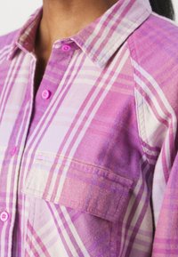 Pink and purple plaid shirt with visible buttons, collared neckline, and front pocket. Made of a textured fabric with varied color tones.