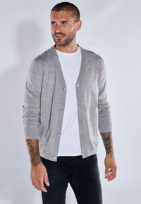 Grauer Cardigan mit V-Ausschnitt, Knopfleiste und langen Ärmeln, aus weichem Stoff, getragen über einem weißen T-Shirt und kombiniert mit dunklen Jeans.