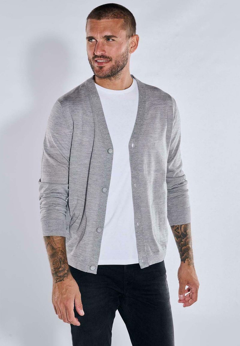 Grauer Cardigan mit V-Ausschnitt, Knopfleiste und langen Ärmeln, aus weichem Stoff, getragen über einem weißen T-Shirt und kombiniert mit dunklen Jeans.