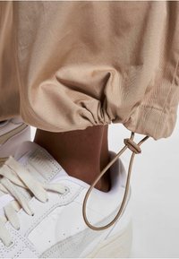Pantaloni bej cu talie elastică, având un tiv elastificat și un șnur maro, asortați cu adidași albi cu suprafețe texturate și șireturi.