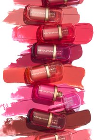 Pequeñas botellas cuadradas de color de labios en varios tonos de rosa y rojo, con acentos dorados y aplicadores, dispuestas sobre muestras de pintura.