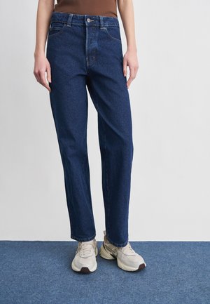 Persona che indossa jeans blu scuro a gamba dritta e sneakers beige, in piedi su un tappeto blu con le mani rilassate lungo i fianchi.