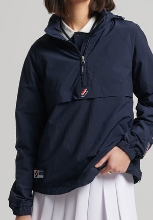 Woman wearing a navy blue half-zip jacket with red and white lightning bolt logo, paired with a white collared shirt and white pleated skirt.