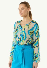 Farbige Bluse mit mutigen blauen, gelben und grünen abstrakten Mustern. Hergestellt aus leichtem Stoff mit lässiger Passform und langen Ärmeln.