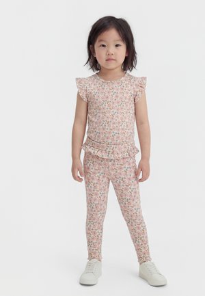 Νεαρή κοπέλα που στέκεται αντικρίζοντας μπροστά, φορώντας ένα ταιριαστό φλοράλ τοπ με βολάν και κολάν, καθώς και άσπρα αθλητικά παπούτσια, σε ένα απλό φόντο.