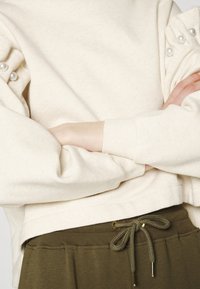 Krémová mikina se perlami na ramenech, spárovaná s olivově zelenými kalhotami se stahovací šňůrkou. Měkká, strukturovaná tkanina.