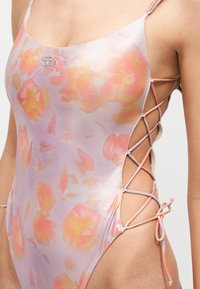 Gros plan d'une femme portant un maillot de bain pastel à motif floral avec des lacets sur les côtés qui laissent apparaître la peau et des bretelles fines.