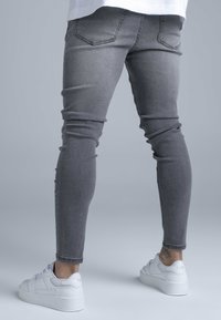 Karcsúsított szürke farmernadrág ötpocketes kialakítással, enyhe kopással és bokáig érő szegéllyel, fehér sportcipővel párosítva.