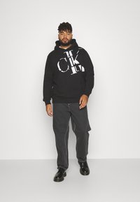 Black hoodie with white Calvin Klein logo, featuring a front pocket and drawstring hood. Worn with dark jeans and black shoes.