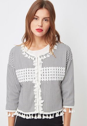 Joven con labios rojos que lleva un cárdigan de cuadros en blanco y negro con borlas blancas en las mangas, el dobladillo y la parte frontal, sobre una blusa blanca.