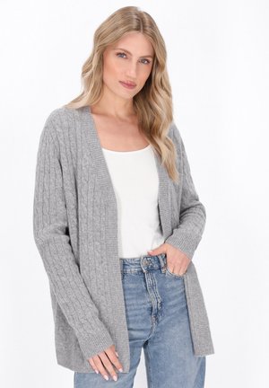 Grijze kabelgebreide cardigan met open voorkant, lange mouwen en geribbelde manchetten, gedragen over een witte top en gecombineerd met high-waisted jeans.