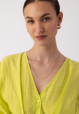 Junge Frau mit zurückgebundenem Haar, trägt goldene Creolen, eine zarte goldene Kettenhalskette und eine leuchtend gelbe, strukturierte V-Ausschnitt-Bluse.