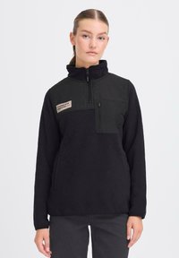 Schwarzer Fleece-Pullover mit hohem Kragen, halbem Reißverschluss, Brusttasche und Seitentaschen; verfügt über ein beiges Etikett auf der oberen linken Seite.
