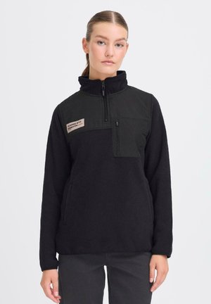 Svart fleece-pull med hög krage, halvdragkedja, bröstficka och sidofickor; har en beige etikett på övre vänstra sidan.
