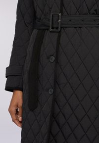 Černý prošívaný kabát s diamantovým vzorem, páskem v pase, knoflíky a kontrastními černými akcenty na límci a manžetách.