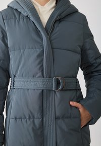 Άτομο που φοράει ένα μπλε επενδυμένο μπουφάν με κουκούλα, με ζώνη στη μέση και ασημένια αγκράφα, ενώ έχει το ένα χέρι στην πλαϊνή τσέπη.