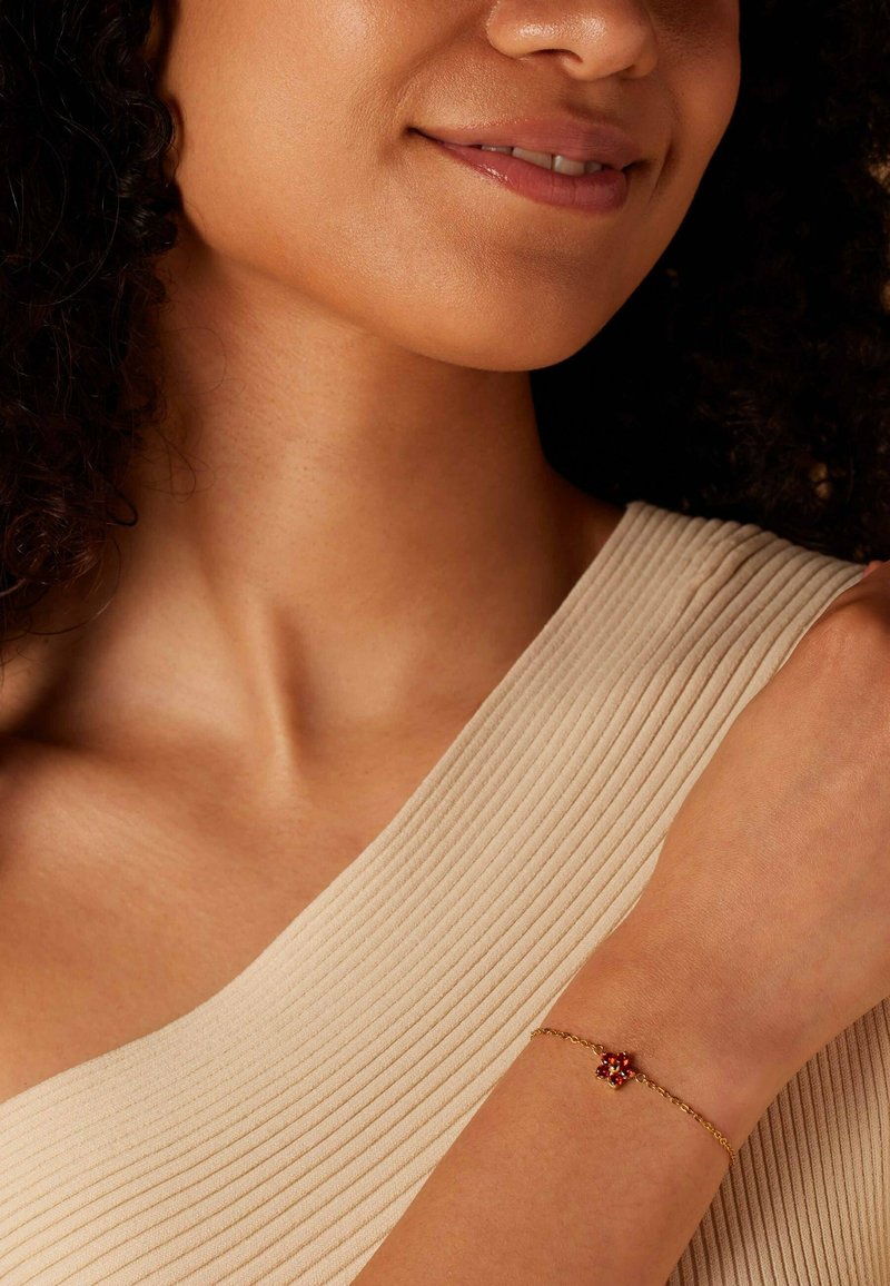 Gouden kettingarmband met een rode edelsteen bloemdetail, gedragen om een pols, tegen een textuur achtergrond van lichtgekleurde stof.