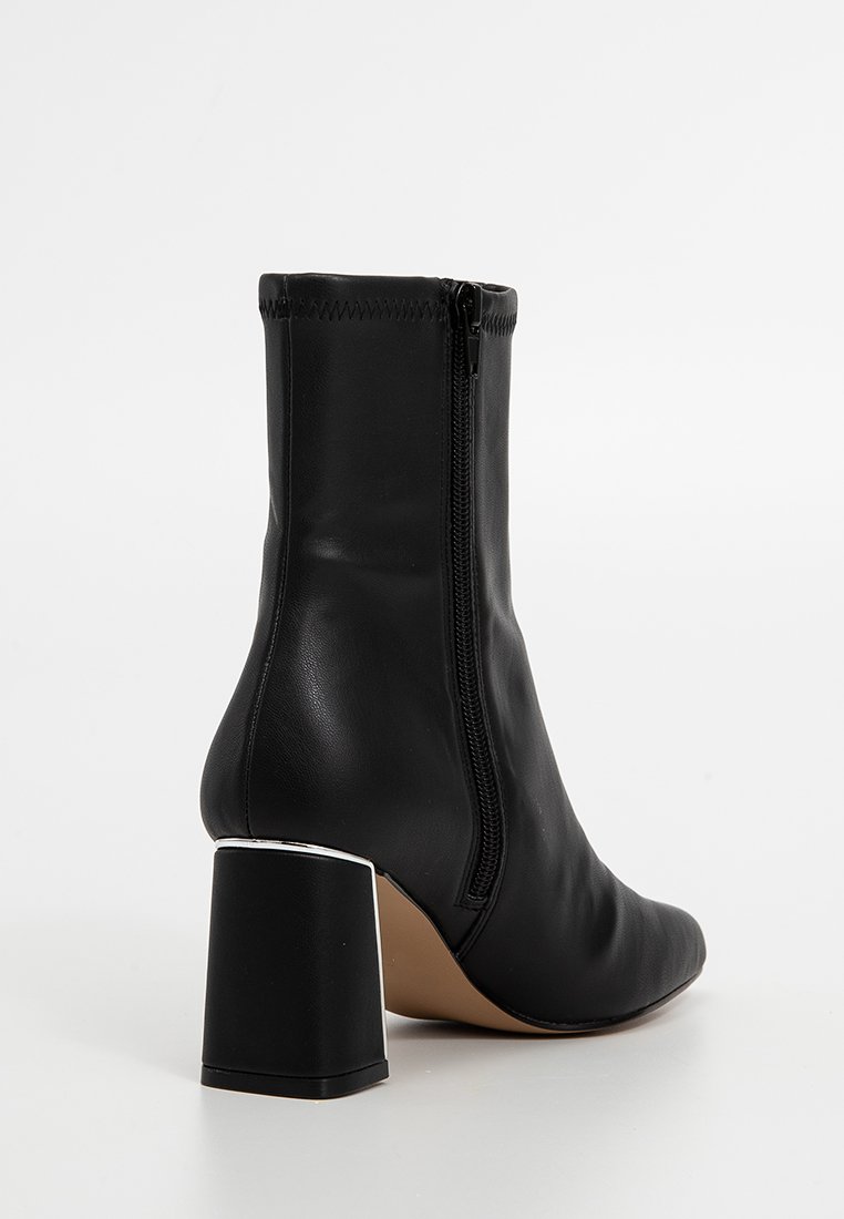 Botín negro de cuero hasta el tobillo con punta redonda, cremallera lateral y un tacón robusto que presenta un acento metálico en la base. Textura suave.