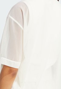 Witte kleding met doorschijnende mouwen, een gladde textuur en een gestructureerd ontwerp met contrasterende biesdetails. De achterkant benadrukt de schoudernaad.