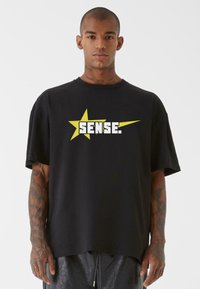 Juodas per didelis T-shirt'as, pagamintas iš minkštos medžiagos, su dideliu geltonu žvaigžde ir baltu tekstu „SENSE.“ priekyje.
