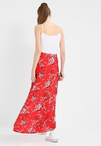 Maxi jupe fleurie rouge au design fluide, présentant des motifs floraux blancs, un tissu léger et une taille élastique. Portée avec un haut blanc.