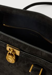Schwarze Wildlederhandtasche mit goldenen Beschlägen, ausgestattet mit einer Reißverschlusstasche und einem charakteristischen Schlossverschluss. Glatte Textur und strukturiertes Design.