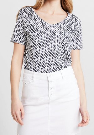 Kobieta w krótkim rękawie, nosząca czarno-białą bluzkę w geometryczny wzór z dekoltem w serek, włożoną w białą spódnicę z guzikami z przodu.