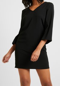 Robe noire en tissu lisse, avec un décolleté en V et des manches trois-quarts, présentant une silhouette ajustée et un détail d fente sur le côté.