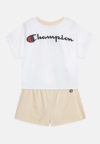 T-shirt bianca in cotone con logo "Champion" nero, abbinata a pantaloni corti beige in cotone con patch del logo sull'orlo. Fit classico e rilassato.