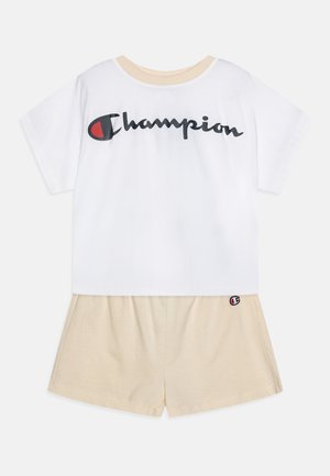 T-shirt bianca in cotone con logo "Champion" nero, abbinata a pantaloni corti beige in cotone con patch del logo sull'orlo. Fit classico e rilassato.