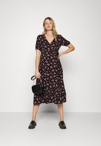 Vestido envolvente preto com padrão floral, de mangas curtas, apresentando uma bainha franzida e pequenas flores em vermelho e branco. Usado com uma bolsa preta e sapatos tipo slip-on.