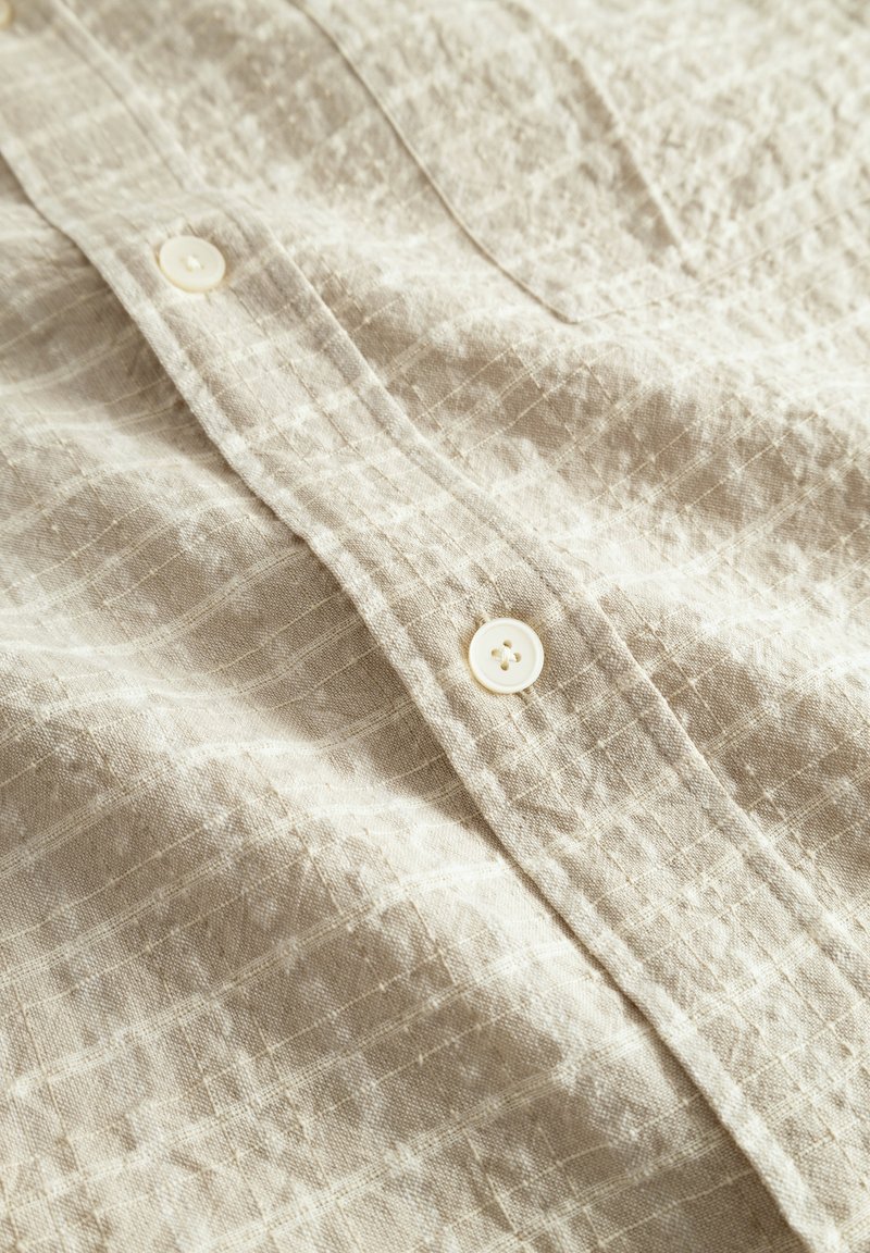 Chemise en lin beige clair avec une surface texturée, dotée de boutons blancs et d'un motif à carreaux sur toute la tissu.