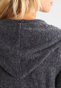 Szara wełniana bluza z kapturem, wykonana z teksturowanej tkaniny, z trójkątnym kapturem i przeszyciami z tyłu. Powierzchnia wydaje się miękka i ciepła.